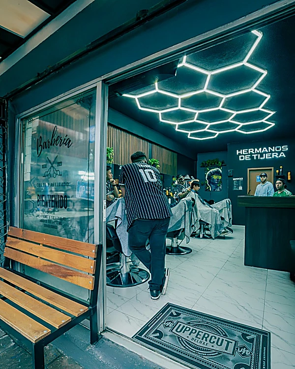 Entrada de Hermanos de Tijera: banca de madera, ventanal con la palabra Barbería y el techo de hexágonos LED encendido sobre las sillas