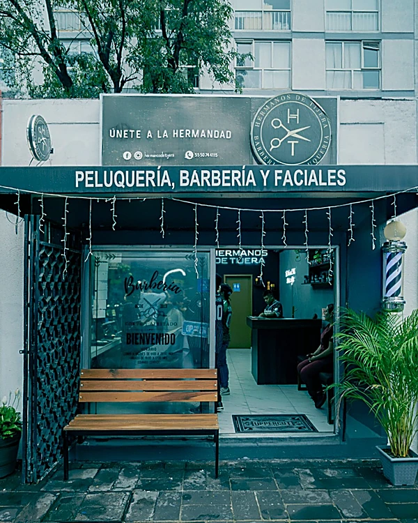 Fachada con el letrero Peluquería, Barbería y Faciales, el lema Únete a la hermandad, banca de madera y poste de barbero