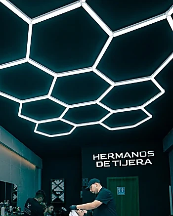 Techo de hexágonos LED y el letrero Hermanos de Tijera al fondo del local, con un barbero trabajando