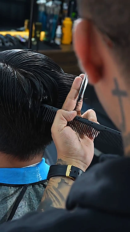Barbero de espaldas, con una cruz tatuada en el cuello, revisando el corte con tijera y peine